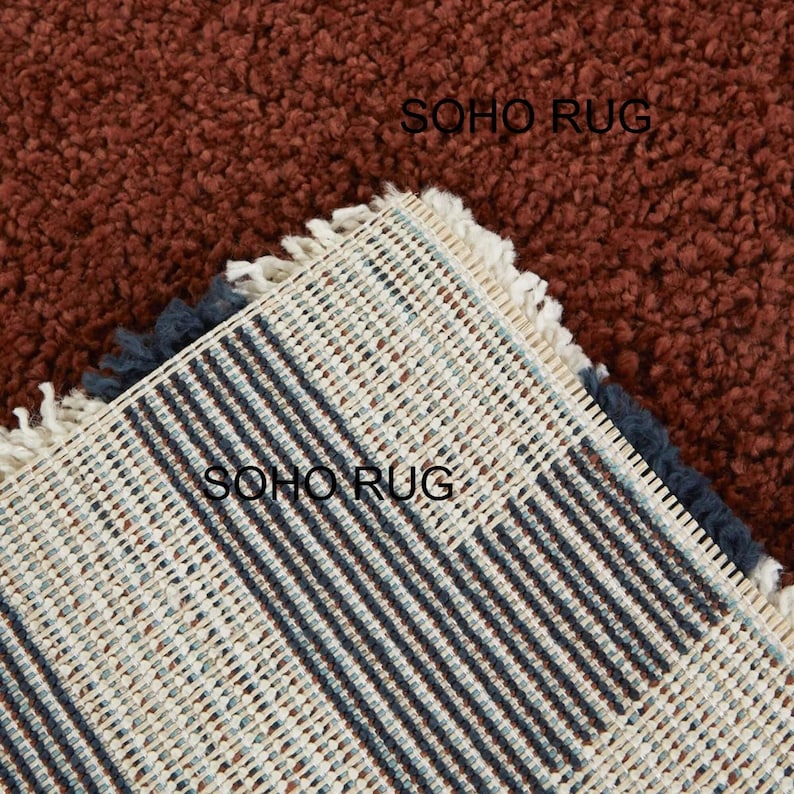 Pu&ograve; includere: Primo piano di un tappeto strutturato color ruggine con un lato inferiore visibile. Il lato inferiore presenta un motivo intrecciato a strisce beige, blu navy e azzurre. Il bordo del tappeto ha una frangia. Il testo "SOHO RUG" &egrave; visibile.