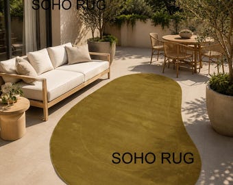May include: An olive green, organically shaped rug with the text "SOHO RUG" in black. The rug is placed on a patio with outdoor furniture, including a sofa, side table, and dining set. The rug's design adds a modern touch to the outdoor space.