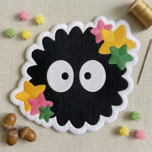 May include: A black embroidered patch featuring a cartoon character with large white eyes and a white scalloped border. The patch is decorated with yellow, pink, and green star-shaped embellishments. Additional decorative elements include acorns, colorful candies, a spool of thread, and a needle.
