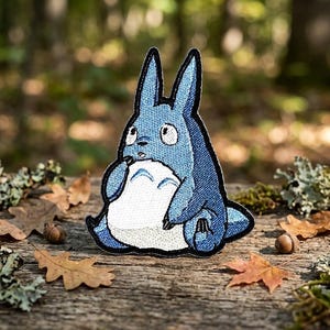 May include: A blue embroidered patch featuring a cartoon character with large ears and a white belly. The patch has a denim-like texture and a black border. The character is sitting down, with a surprised expression. Acorns and leaves surround the patch.