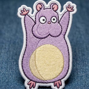May include: Embroidered patch featuring a cartoon hamster in shades of purple, yellow, and pink. The hamster has its arms raised and a large belly. The patch is outlined in white.