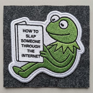 May include: Embroidered patch featuring a green frog reading a book titled "HOW TO SLAP SOMEONE THROUGH THE INTERNET." The frog is outlined in white, set against a gray felt background. A humorous, pop culture-themed item.