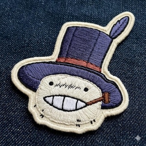 May include: Embroidered patch featuring a smiling character wearing a purple top hat with a feather, a brown band, and a cigar. The character has a white face with a wide grin and small black eyes. The patch is outlined in cream.