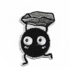 May include: Embroidered patch featuring a cartoon character with a chef's hat. The character is a black, round figure with large white eyes, rosy cheeks, and small legs, giving the impression of movement. The patch has a white border.