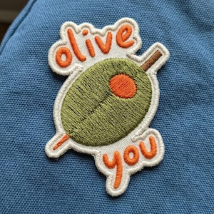 May include: Embroidered patch featuring a green olive with an orange pit and a toothpick. The patch has the words "olive you" in orange script. The patch is outlined in white and is on a blue background.
