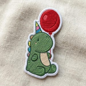 May include: Embroidered patch featuring a green dinosaur wearing a party hat and holding a red balloon. The dinosaur has colorful spines and a tan belly. The patch is outlined in white.