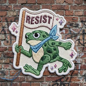 May include: Embroidered patch featuring a green frog wearing a blue bandana and holding a flag that reads "RESIST" in maroon letters. The patch has a white border with embroidered star accents and is set against a brick wall.
