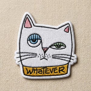 May include: Embroidered white cat patch with a whimsical design. The cat has one blue eye and one green eye, pink inner ears, and a pink nose. A yellow band at the bottom reads "WHATEVER" in black letters. The patch is outlined in black.