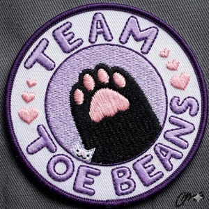 May include: Embroidered patch with a purple border and the words "TEAM TOE BEANS" in purple. The patch features a black cat paw with pink pads, a small white cat, and pink hearts.