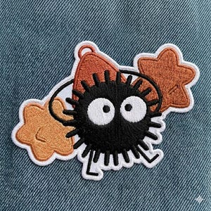 May include: Embroidered patch featuring a black soot sprite with large white eyes, surrounded by orange star and bow-shaped elements, all outlined in white. The patch is set against a blue denim background.