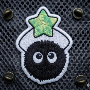 May include: Embroidered patch featuring a black, fluffy creature with large, white-rimmed eyes and a green star on top. The patch is outlined in white and set against a gray mesh background with four bronze-colored fasteners.