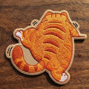 May include: Embroidered patch featuring a cartoon cat from the rear, with orange and brown stripes. The cat is in a playful pose, with its tail and paws visible. The patch has a tan border and is set against a wooden background.