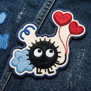 May include: Embroidered patch featuring a black, round character with large white eyes and a spiky body. Two red heart-shaped balloons are attached. The patch has a white background with blue and white accents, set against a denim background.