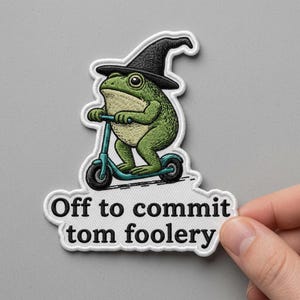 May include: Embroidered patch featuring a green frog wearing a black witch hat and riding a teal scooter. The patch has the text "Off to commit tom foolery" in black letters.