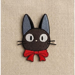 May include: Embroidered patch featuring a black cat with large white eyes, gray ears, and a red bow tie. The patch is centered on a beige fabric background, showcasing the detailed stitching and vibrant colors of the cat design.