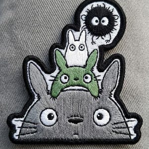 May include: Embroidered patch featuring characters from the animated film, My Neighbor Totoro. The patch includes a large gray Totoro, a green Totoro, a white Totoro, and a Soot Sprite, all with black outlines.
