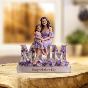 May include: A decorative figurine featuring a mother and child in lavender dresses, seated atop the word "MUM". The letters are filled with purple irises and surrounded by floral accents. The base reads "Happy Mother's Day".