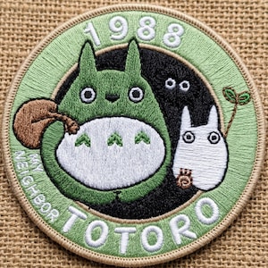 May include: Embroidered patch featuring characters from "My Neighbor Totoro." The design includes a green Totoro, a white Totoro, and the text "1988" and "My Neighbor Totoro" in white. The patch has a green border.