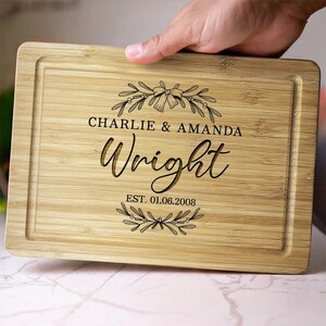 May include: A rectangular bamboo cutting board with a juice groove around the edge. The board is engraved with the names "Charlie & Amanda Wright" and the date "EST. 01.06.2008", along with floral embellishments. The board is held by a hand.