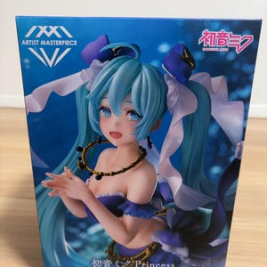 以下が含まれることがあります： 初音ミクプリンセスフィギュアの箱。フィギュアは長い水色の髪で、青と紫のドレスを着ています。箱には「ARTIST MASTERPIECE」の文字と日本語のテキストがあります。