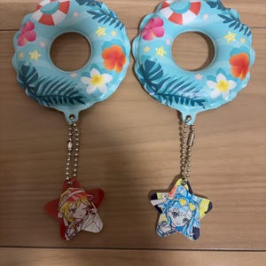 以下が含まれることがあります： 水色の浮き輪のデザインのキーホルダー2つ。それぞれに花柄と葉柄が施されています。各キーホルダーには、漫画のキャラクターのイラストが描かれた星型のチャームが付いており、銀色のチェーンでリングに接続されています。