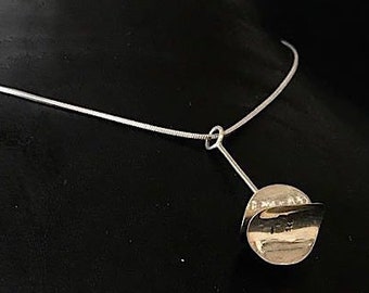 May include: A silver necklace with a circular pendant. The pendant has a curved, metallic design and is attached to the chain by a small silver bar. The necklace is displayed on a black mannequin.