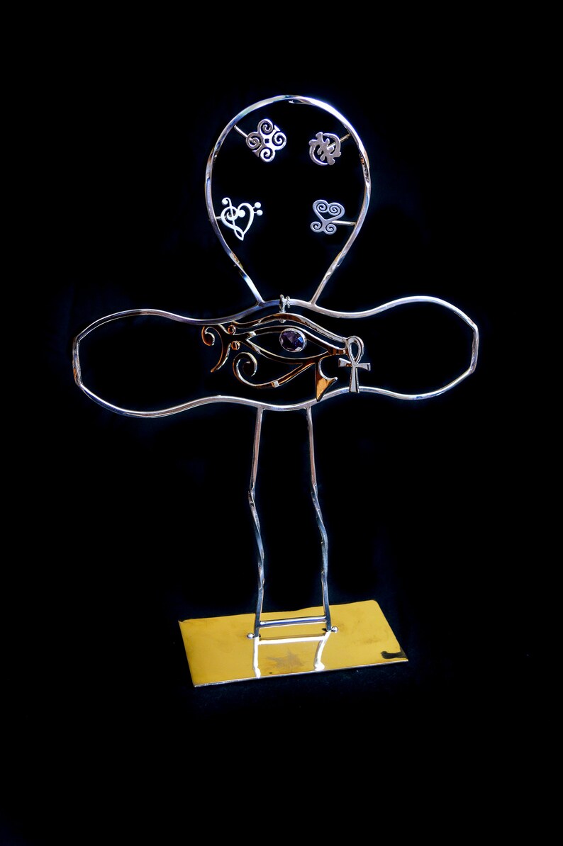 K&ouml;nnte beinhalten: Ein silberner Ankh-St&auml;nder aus Metall mit einem violetten Stein-Horusauge und einem goldenen Ankh-Anh&auml;nger. Der St&auml;nder hat eine goldene Basis und ist mit kleinen silbernen Anh&auml;ngern verziert.