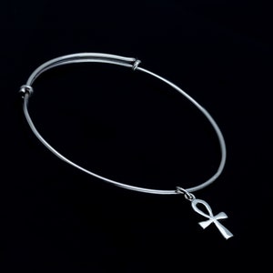 K&ouml;nnte beinhalten: Ein silbernes, verstellbares Armreif-Armband mit einem silbernen Ankh-Anh&auml;nger. Das Armband befindet sich auf einem schwarzen Hintergrund.