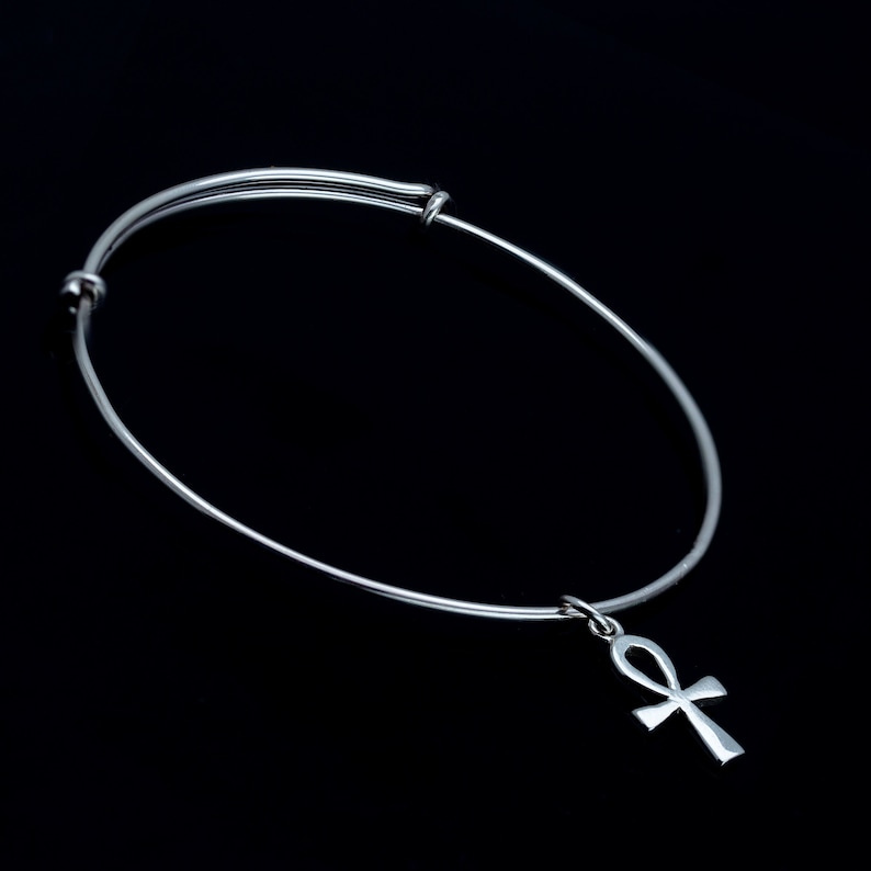 K&ouml;nnte beinhalten: Ein silbernes, verstellbares Armreif-Armband mit einem silbernen Ankh-Anh&auml;nger. Das Armband befindet sich auf einem schwarzen Hintergrund.
