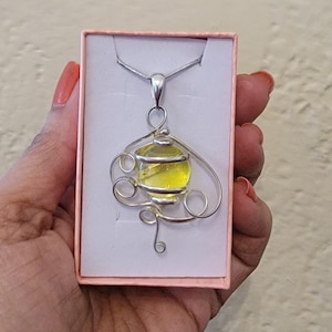 May include: A silver pendant necklace featuring a yellow orb encased in a decorative wire frame. The pendant is displayed in a pink jewelry box. The necklace has a delicate silver chain.