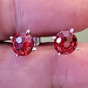 May include: A pair of stud earrings featuring round, faceted red gemstones set in a silver metal. The earrings are displayed against a blurred background, showcasing the vibrant color and sparkle of the gems.