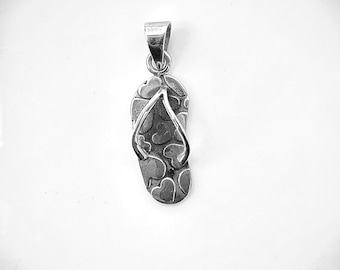May include: A silver-colored pendant in the shape of a flip-flop. The flip-flop has a heart pattern on the sole and a metal strap. The pendant is designed to be worn as a necklace or charm.