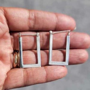 May include: A pair of silver hoop earrings with a rectangular shape. The earrings are simple and elegant.