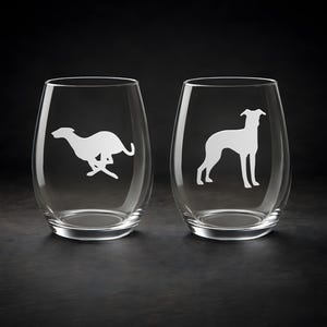 May include: Two stemless wine glasses with etched white greyhound dog designs. One glass shows a running greyhound, the other a standing greyhound. The glasses are clear and sit on a dark surface.