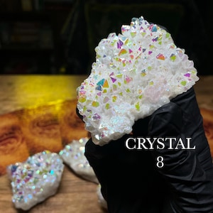 May include: A cluster of white crystals with iridescent, rainbow-colored facets. The crystals are held by a gloved hand. The text "CRYSTAL 8" is visible. Other crystals are visible in the background.