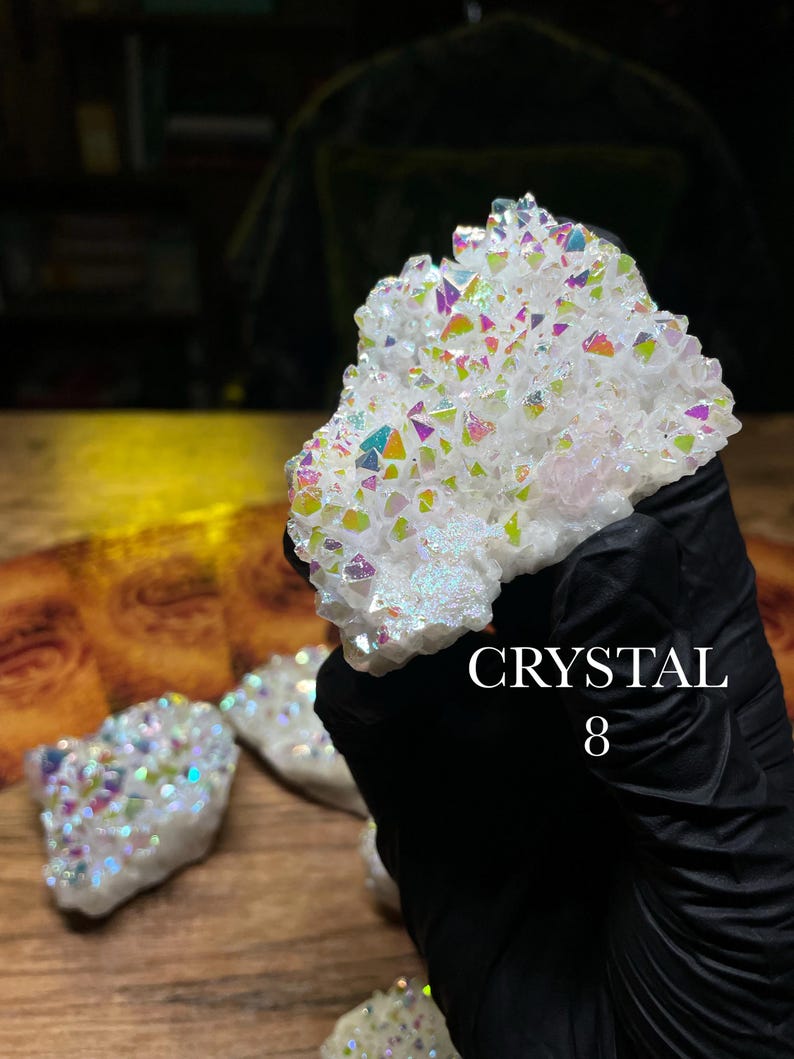 May include: A cluster of white crystals with iridescent, rainbow-colored facets. The crystals are held by a gloved hand. The text "CRYSTAL 8" is visible. Other crystals are visible in the background.