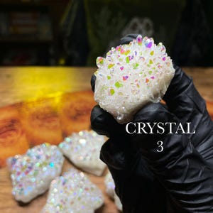 May include: A cluster of white crystals with iridescent rainbow hues. The crystals are held in a gloved hand, with several other similar crystals visible on a wooden surface. The text "CRYSTAL 3" is displayed.