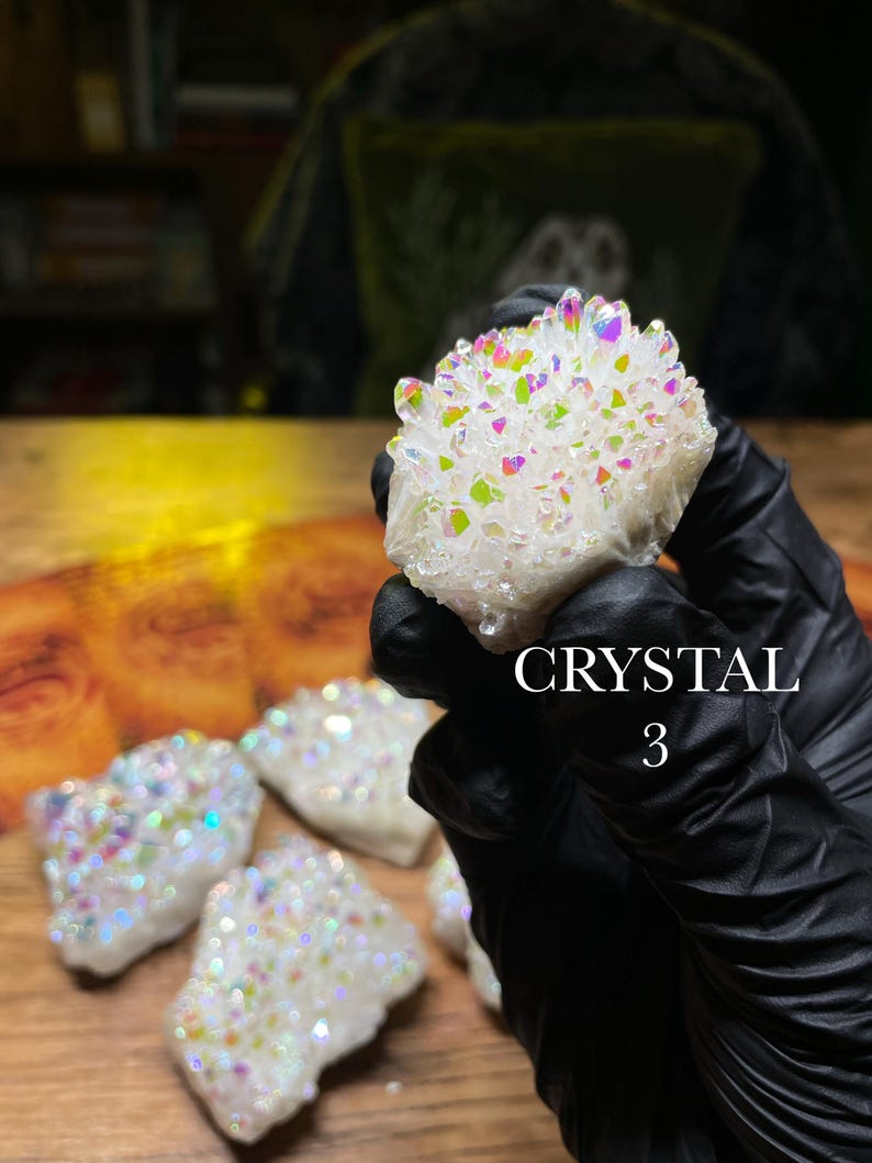 May include: A cluster of white crystals with iridescent rainbow hues. The crystals are held in a gloved hand, with several other similar crystals visible on a wooden surface. The text "CRYSTAL 3" is displayed.