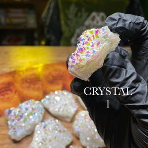 May include: A collection of white crystals with iridescent, rainbow-colored surfaces. The crystals are held by a gloved hand, with the text "CRYSTAL 1" visible. The crystals are displayed on a wooden surface.