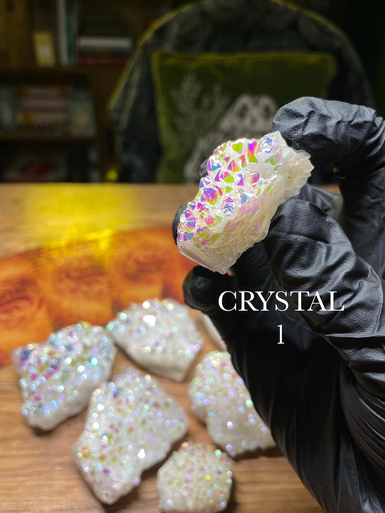 May include: A collection of white crystals with iridescent, rainbow-colored surfaces. The crystals are held by a gloved hand, with the text "CRYSTAL 1" visible. The crystals are displayed on a wooden surface.