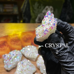 May include: A collection of white crystals with iridescent, rainbow-colored coatings. The crystals are held by a gloved hand, with the text "CRYSTAL 2" visible. The crystals are displayed on a wooden surface.