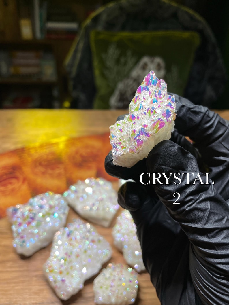 May include: A collection of white crystals with iridescent, rainbow-colored coatings. The crystals are held by a gloved hand, with the text "CRYSTAL 2" visible. The crystals are displayed on a wooden surface.