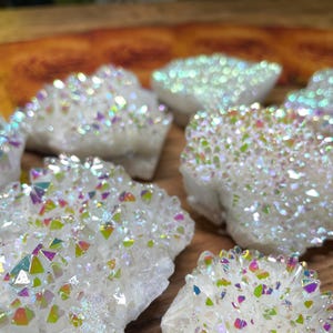 May include: Close-up of several white aura quartz clusters. The crystals have a sparkling, iridescent coating that reflects a rainbow of colors. The clusters are arranged on a wooden surface, with some in focus and others blurred.