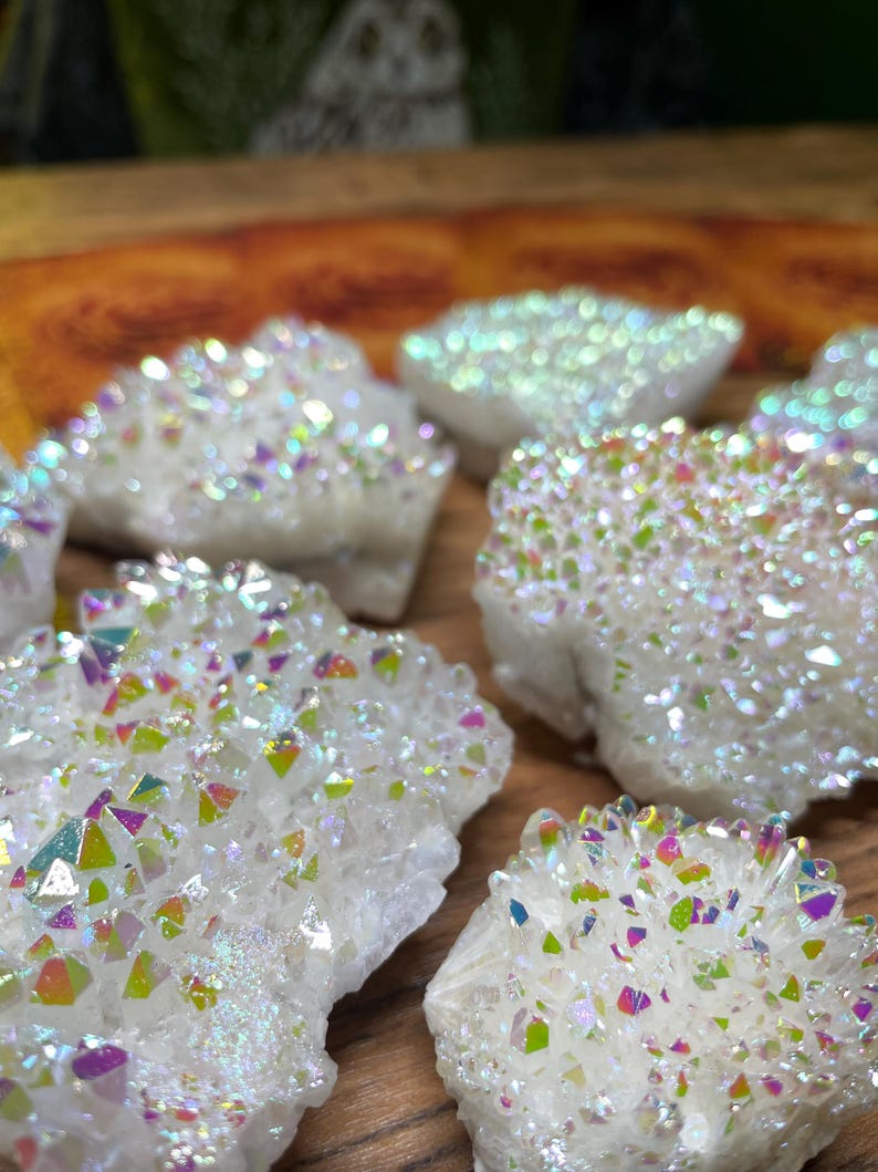 May include: Close-up of several white aura quartz clusters. The crystals have a sparkling, iridescent coating that reflects a rainbow of colors. The clusters are arranged on a wooden surface, with some in focus and others blurred.