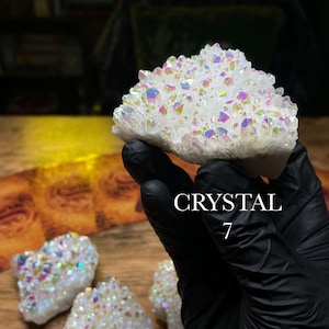May include: A cluster of white crystals with iridescent rainbow hues. The crystals are held by a gloved hand, with the word "CRYSTAL" and the number "7" in the image. Several other crystals are visible on a wooden surface.