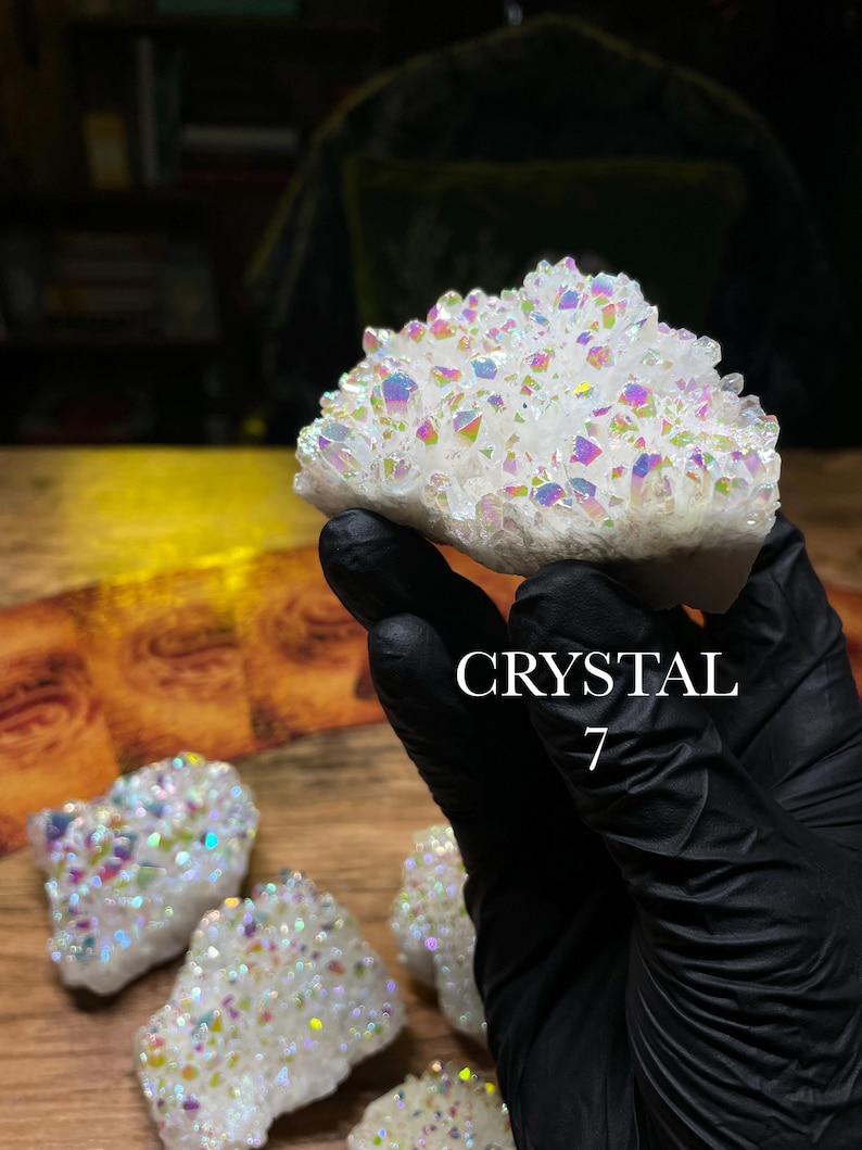 May include: A cluster of white crystals with iridescent rainbow hues. The crystals are held by a gloved hand, with the word "CRYSTAL" and the number "7" in the image. Several other crystals are visible on a wooden surface.