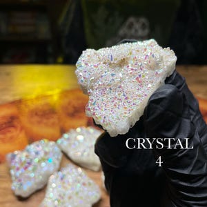 May include: A cluster of iridescent crystals with a white base and colorful, shimmering surfaces. The crystals are held by a gloved hand. The text "CRYSTAL 4" is visible in the image. Several other crystals are visible in the background.