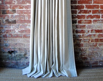May include: White linen curtains with a blue and white geometric pattern border. The curtains are hanging from a black curtain rod against a red brick wall.