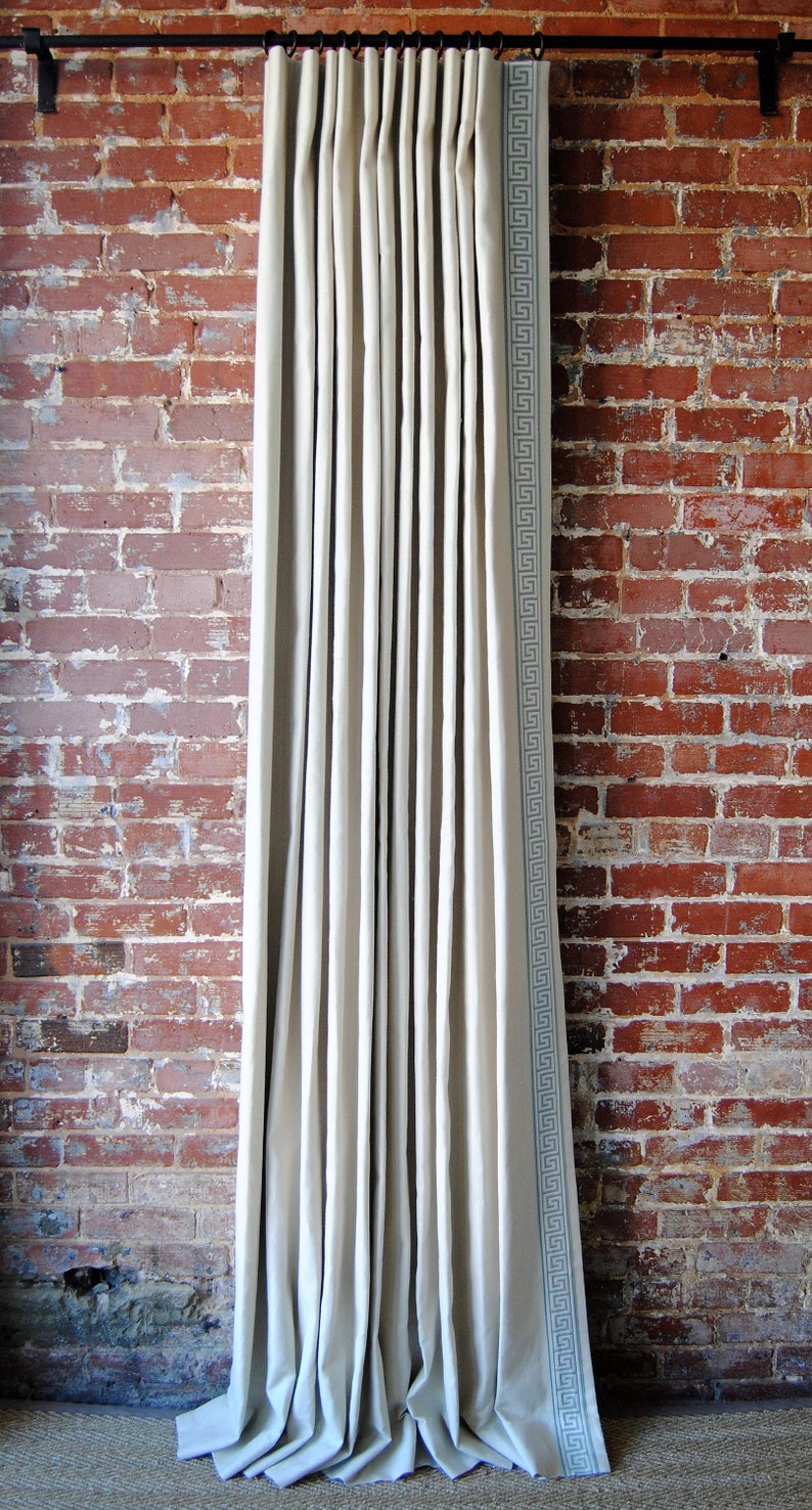 May include: White linen curtains with a blue and white geometric pattern border. The curtains are hanging from a black curtain rod against a red brick wall.