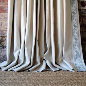 May include: A pair of white linen curtains with a gray Greek key trim hanging in front of a brick wall.